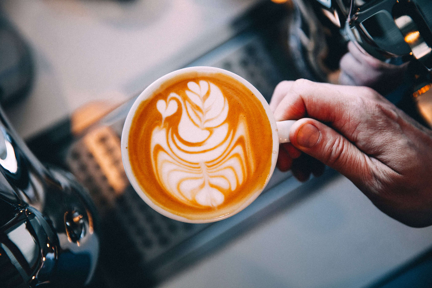 Hand holding a cup of Kirenge Gold Kenyan premium coffee with latte art, rich flavor, blurred café background.