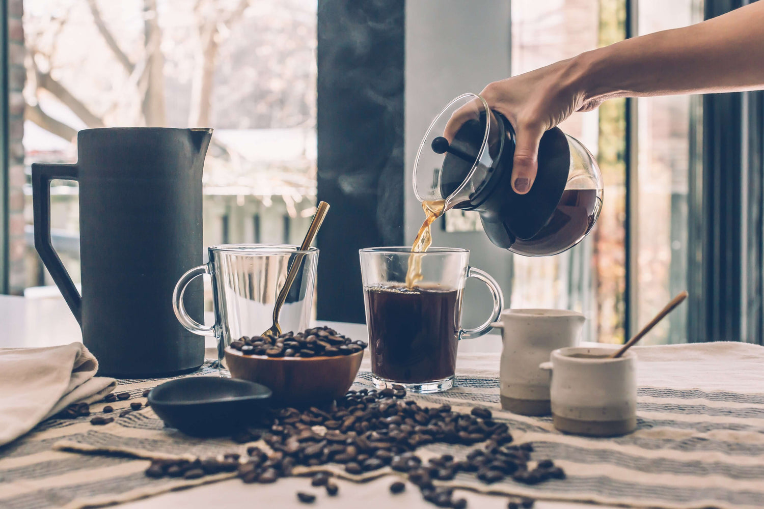 Person pouring Kirenge Gold coffee from a glass carafe into a glass mug on a table with coffee beans and a coffee filter.