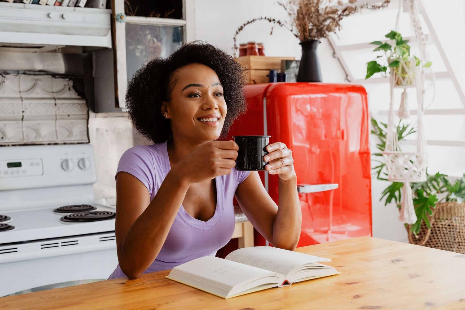 Woman sipping Kenyan premium coffee from Kirenge Gold mug, cozy kitchen scene with red fridge and book on table.