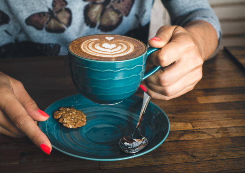 Person holding a blue mug with a cappuccino and cookies on a wooden table
